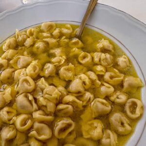 Bowl of cheese tortellini in clear broth with a spoon, and a wine glass in the background.