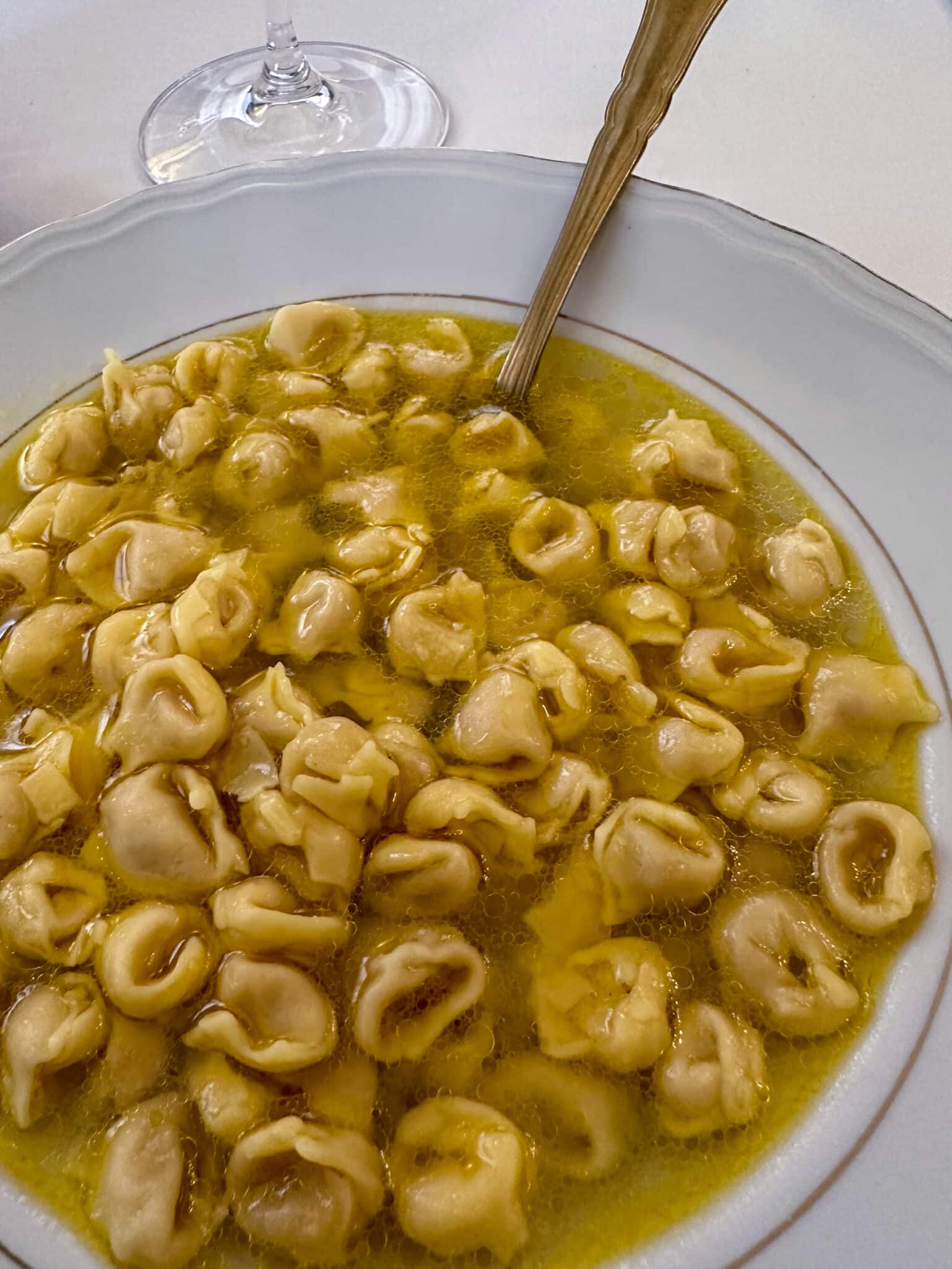 Bowl of cheese tortellini in clear broth with a spoon, and a wine glass in the background.